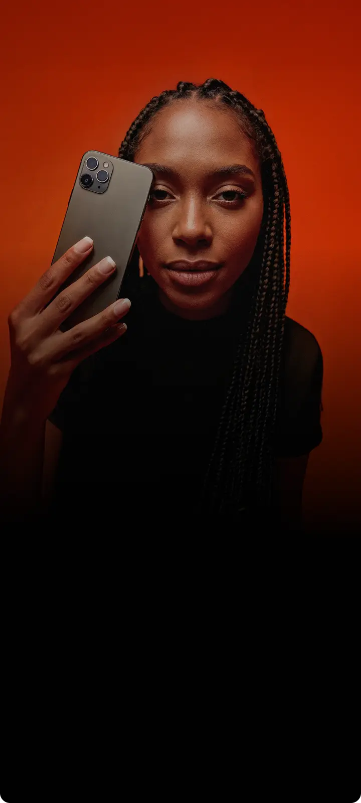 No centro da imagem há uma jovem mulher com pele escura e cabelo com tranças, usando uma blusa preta, segurando um smartphone cinza na frente do rosto, com expressão neutra e fundo laranja e marrom em degradê